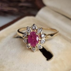 May include: A gold ring featuring an oval ruby gemstone at its centre, encircled by a halo of small, clear diamonds. The ring has a floral or starburst design. It is presented in a vintage jewellery box.
