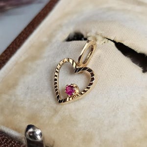 Peut inclure: Un pendentif en forme de cœur en or avec une petite pierre précieuse rouge et ronde au centre. Le pendentif est présenté à l'intérieur d'un écrin à bijoux vintage avec un intérieur de couleur crème. Le cœur a un motif texturé sur les bords.