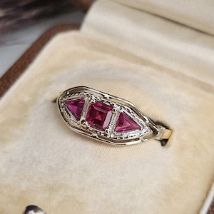 May include: A vintage-style ring with a gold band and a silver-tone setting. The ring features three vibrant, square and triangular, red gemstones. The ring is displayed inside a brown jewelry box with a beige interior.
