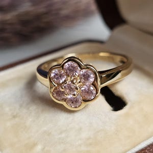May include: A gold ring featuring a flower design with pink gemstones. The ring is set in a gold band and is displayed inside a brown jewelry box with a beige interior.