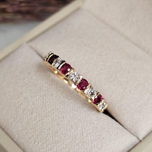 Könnte beinhalten: Ein goldener Bandring mit abwechselnd roten Edelsteinen und klaren, funkelnden Steinen. Der Ring ist in einem goldenen Band gefasst und wird in einer hellbeigen Ringschatulle präsentiert. Die Edelsteine sind gleichmäßig um den Ring herum angeordnet.