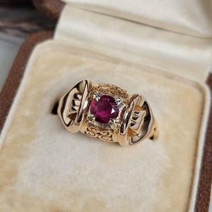 May include: A gold ring featuring a large, oval, ruby-coloured gemstone at its centre. The ring has an elaborate design with filigree and decorative features. It is presented in a vintage jewellery box with a cream-coloured interior.