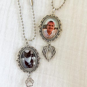May include: Two silver-toned memorial necklaces. One features a photo of a person in an oval frame with a wing charm. The other has a photo of a black and white cat in an oval frame with a paw print charm. Both hang on ball chains.