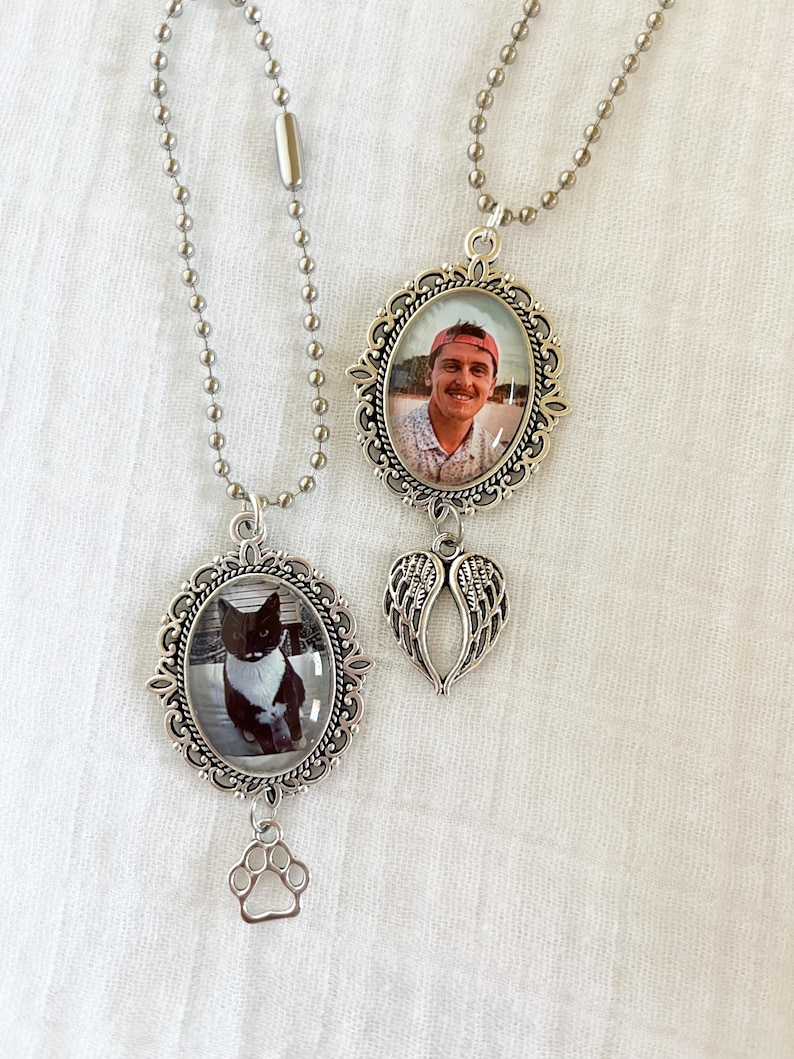 May include: Two silver-toned memorial necklaces. One features a photo of a person in an oval frame with a wing charm. The other has a photo of a black and white cat in an oval frame with a paw print charm. Both hang on ball chains.