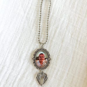 May include: A silver-toned memorial photo pendant with a ball chain. The pendant features an oval frame with a photo of a person wearing a red cap. Below the frame hangs a pair of silver wings.