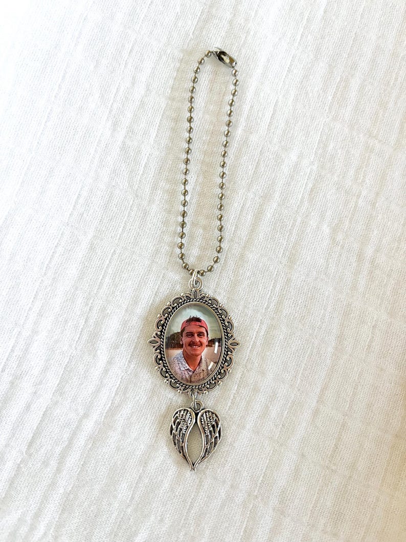 May include: A silver-toned memorial photo pendant with a ball chain. The pendant features an oval frame with a photo of a person wearing a red cap. Below the frame hangs a pair of silver wings.