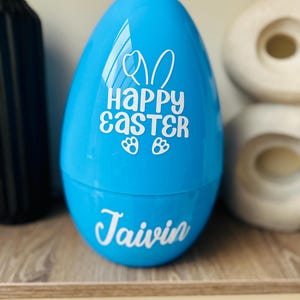 May include: A vibrant blue egg-shaped container, featuring the words "Happy Easter" and a bunny design in white. The name "Taivin" is also written in white on the egg. The container is placed on a wooden shelf.