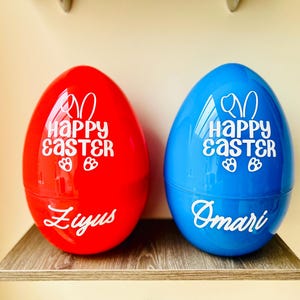 May include: Two large, plastic Easter egg containers, one red and one blue, sit on a wooden shelf. Each egg has the words "Happy Easter" in white, along with bunny ears and paw prints. The red egg says "Ziyus" and the blue egg says "Omari".