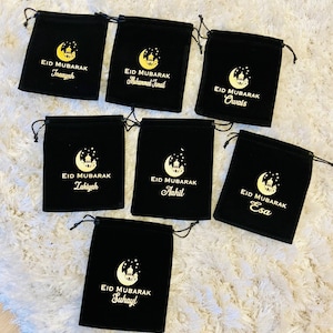 May include: Seven black velvet drawstring bags with gold embroidery. Each bag has a crescent moon and a mosque design with the text "Eid Mubarak" and a name. The names are Inaayah, Muhammed Ismail, Owais, Zakiyah, Aahil, Esa, and Suhayl.