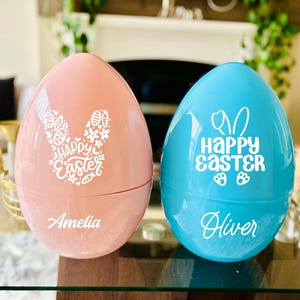 May include: Two large, egg-shaped containers, one pink and one blue, with white text and graphics. The pink egg has a bunny design and the words "Happy Easter" and the name "Amelia." The blue egg has the words "Happy Easter" and the name "Oliver."
