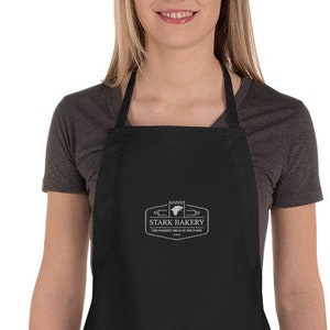 Puede incluir: Delantal negro con un logotipo bordado en blanco que dice "Stark Bakery" y un pequeño gráfico de un rodillo y una hogaza de pan.