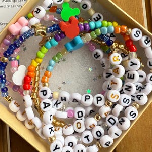 Könnte beinhalten: Eine Sammlung farbenfroher Perlenarmbänder, darunter solche mit Buchstaben, die Wörter wie "HAPPY" und "YOU" buchstabieren. Die Armbänder befinden sich in einer kleinen Schachtel mit einer Pflegeanleitung.