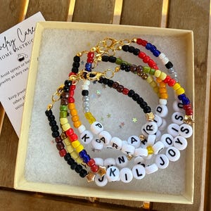 May include: A collection of beaded bracelets in a light yellow box. The bracelets feature colorful beads and white letter beads spelling out names. Gold-colored clasps and star charms are also visible. A card with "Jewelry Care" instructions is in the box.