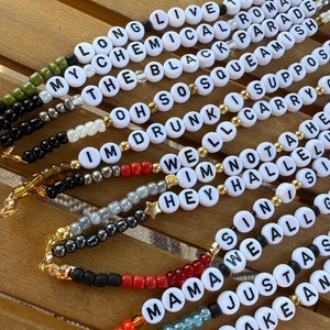 Könnte beinhalten: Nahaufnahme von zehn bunten Perlenarmbändern mit weißen Buchstabenperlen, die Songtitel und Songtexte buchstabieren. Die Armbänder liegen auf einer Holzoberfläche.