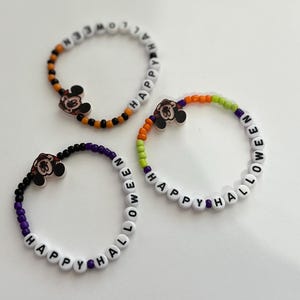 Puede incluir: Tres pulseras de cuentas con dijes de Mickey Mouse. Las pulseras deletrean "Happy Halloween" en letras blancas sobre cuentas negras, naranjas y verdes.
