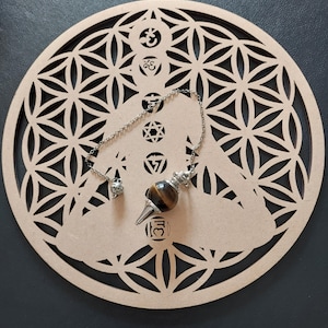 May include: A brown and gold tiger's eye pendulum hanging from a silver chain on a wooden base with a flower of life design. The base also features symbols like the Star of David and Om.