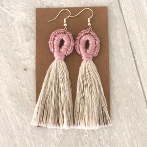 Può includere: Un paio di orecchini a nappa rosa e crema. Gli orecchini sono realizzati con un cerchio rosa all'uncinetto e una lunga nappa crema.