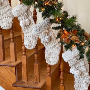 May include: Four cream-colored crocheted Christmas stockings with white faux fur trim and pom-poms hanging on a wooden staircase railing decorated with a garland of green pine branches and gold ornaments.