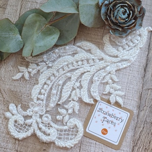 May include: White lace applique with pearl embellishments. The applique has a floral design with a leaf pattern. The applique is on a wooden surface with a tag that says "Strawberry Taces Etsy".