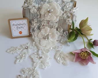 May include: White beaded floral lace trim with pearl and crystal embellishments. The trim is displayed on a metal container and next to a pink flower.