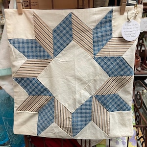 May include: A square pillow cover featuring a central star pattern made of cream, blue plaid, and brown striped fabric. A white circular tag attached to the top right corner reads: Pillow cover for throw pillow Measures 14"x14" Needs pillow form $25.00.
