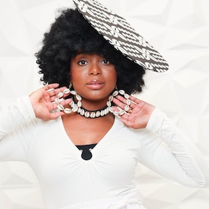 May include: A woman wearing a white long-sleeved shirt, a white skirt with a bow, and a large black and white patterned hat. She is also wearing a white shell necklace and earrings.