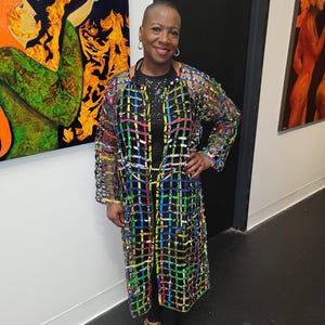 May include: A woman wearing a long, open-front coat with a colorful grid pattern over a black outfit. The coat features a vibrant mix of colors, including blue, yellow, green, and pink. She is wearing leopard print shoes and hoop earrings. Artwork hangs on the wall behind her.