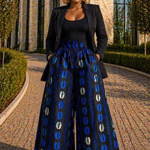 May include: A woman wearing a black blazer over a black top and wide-leg pants. The pants are black with a blue and white oval pattern. The woman has short blonde hair and is standing on a brick pathway.