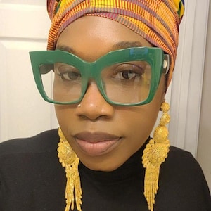 May include: A person wearing a colorful head wrap, green cat-eye glasses, and long gold dangle earrings. The earrings have a round, textured design at the top and a long, chain-like fringe.