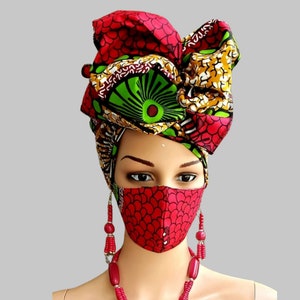 May include: A mannequin head adorned with a vibrant African print head wrap, matching face mask, and beaded jewelry. The head wrap features red, green, yellow, and white patterns. The mask and earrings are red with a scale-like design. The necklace is red.
