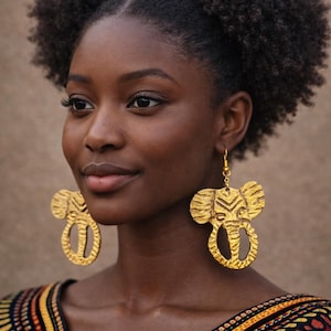 May include: A woman wearing gold-colored elephant-shaped earrings. The earrings have a textured surface and are suspended from gold hooks. She is wearing a colorful patterned top with red, yellow, and black details. Her hair is styled in two buns.