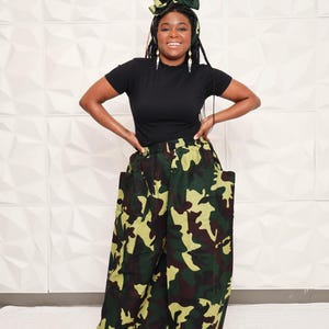 May include: A black short-sleeved top paired with wide-leg camouflage pants. The pants feature a green, brown, and tan pattern. A matching camouflage bow-shaped head wrap completes the outfit. The model is smiling.