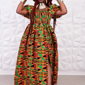 May include: A vibrant, patterned maxi dress with a matching hat. The dress features a high slit and short sleeves. The color palette includes orange, green, black, and yellow. Gold jewelry accents the outfit.