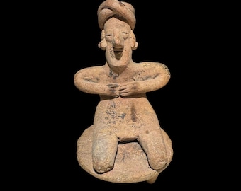 May include: A small, ancient terracotta figurine of a seated figure with arms crossed over the chest. The figure has a turban-like headdress and a smiling expression. The sculpture is a light brown color and sits on a circular base.