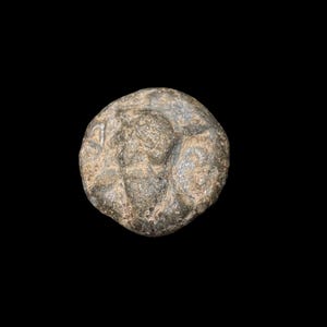 May include: A weathered, circular coin with a detailed relief. The coin's surface is a mix of gray and brown tones, with visible signs of age and wear. The relief depicts a human figure and other design elements.