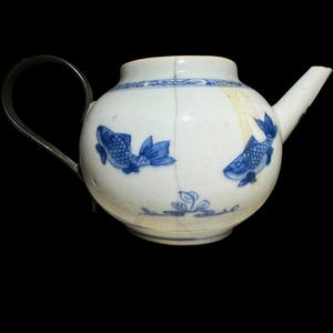May include: White ceramic teapot with blue fish and floral patterns. It features a silver handle and spout. The round teapot has a crack down the side. The teapot is set against a black backdrop.