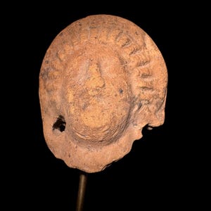 Puede incluir: Una escultura de terracota de una cabeza, posiblemente antigua. La cabeza es de color marrón claro con detalles tallados que sugieren cabello. Se puede ver un agujero en el lateral. La escultura está montada sobre una varilla metálica sobre un fondo negro.