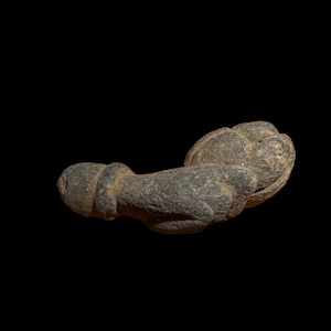 May include: A weathered, gray stone sculpture of an abstract form. The sculpture has a bulbous, rounded shape on one end and a cylindrical shape on the other, with a textured surface. The object is set against a black background.