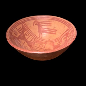 Puede incluir: Un cuenco de cerámica de color terracota con un pájaro rojo y diseños geométricos. El pájaro está representado con un cuello largo y alas extendidas. El borde del cuenco presenta patrones repetidos y el fondo es negro.