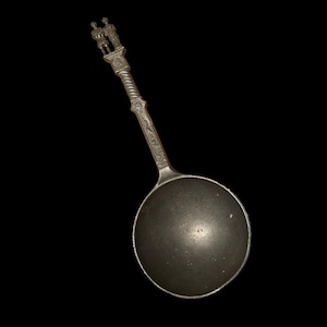Puede incluir: Cuchara antigua de color plateado con un cuenco redondo y un mango detallado. El mango presenta un diseño retorcido y una parte superior decorativa con dos pequeñas figuras. La cuchara está sobre un fondo negro.