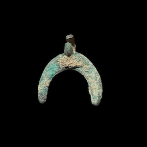 May include: An ancient bronze artefact with a curved, horseshoe-like form. It features a small, raised projection at the top and displays signs of age, with a greenish patina and textured surface. The background is a solid black.