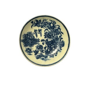 Può includere: Un piccolo piatto rotondo in ceramica antica con una base color crema e un motivo floreale blu. Il design include fiori, alberi e un piccolo edificio. Il piatto ha un bordo leggermente rialzato.