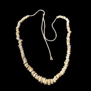 Puede incluir: Un collar compuesto por pequeñas cuentas irregulares, de color blanquecino, ensartadas en un cordón color crema. Las cuentas tienen un aspecto natural y orgánico, y el cordón está anudado en los extremos. El fondo es negro liso.