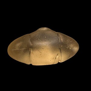 Peut inclure: Un objet doré de forme ovale, avec un sommet arrondi et trois indentations verticales. L'objet a une surface lisse, légèrement texturée, sur un fond noir. La forme générale ressemble à une petite soucoupe volante stylisée.