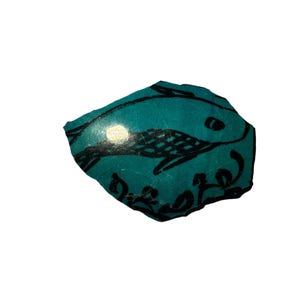Puede incluir: Una piedra de color turquesa, de forma irregular, con un diseño de pez negro. El pez está estilizado con un cuerpo estampado y una cola curvada. La piedra tiene un acabado brillante y parece hecha a mano.