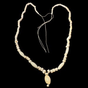 Könnte beinhalten: Beigefarbene Perlenkette mit einem zentralen Anhänger. Die Halskette besteht aus verschiedenen Perlenformen, darunter zylindrische und ovale, die mit einer dünnen, beigefarbenen Schnur aufgefädelt sind. Der Anhänger hat eine ovale Form mit einer kleinen, runden Perle unten.