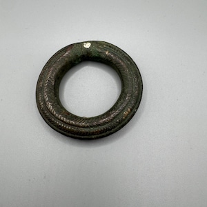 Può includere: Un anello antico in bronzo di forma circolare con un motivo intrecciato e strutturato. L'anello presenta una patina scura e invecchiata con sfumature verdi e marroni, che indicano l'età e l'uso. La superficie dell'anello mostra segni di usura.