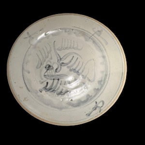 May include: A circular, off-white ceramic plate with a hand-painted blue design. The design features a bird-like figure in the centre, surrounded by floral or abstract patterns. The plate has a slightly raised rim against a black background.