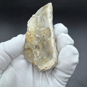 May include: A pale, translucent flint stone with a smooth, chipped surface featuring variegated shades of cream, light gray, and tan. The stone has an elongated, irregular shape with sharp, defined edges and a natural, weathered texture throughout.