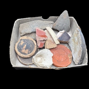 May include: A collection of assorted ceramic and stone fragments contained within a square cardboard tray. The pieces include circular pottery bases in black and orange, jagged grey stone shards, a red clay fragment, and various textured white pieces.